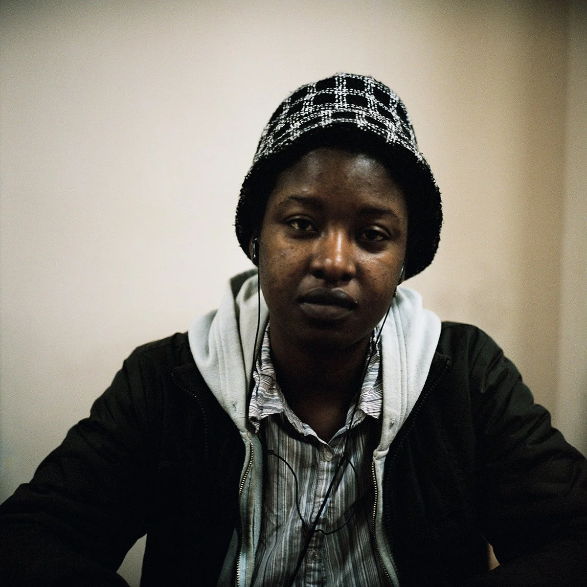 Une jeune femme portant un bonnet à carreaux noirs et blancs, une veste noire, un sweat à capuche gris, une chemise à rayures et des écouteurs, assise face à la caméra dans une pièce aux murs clairs.