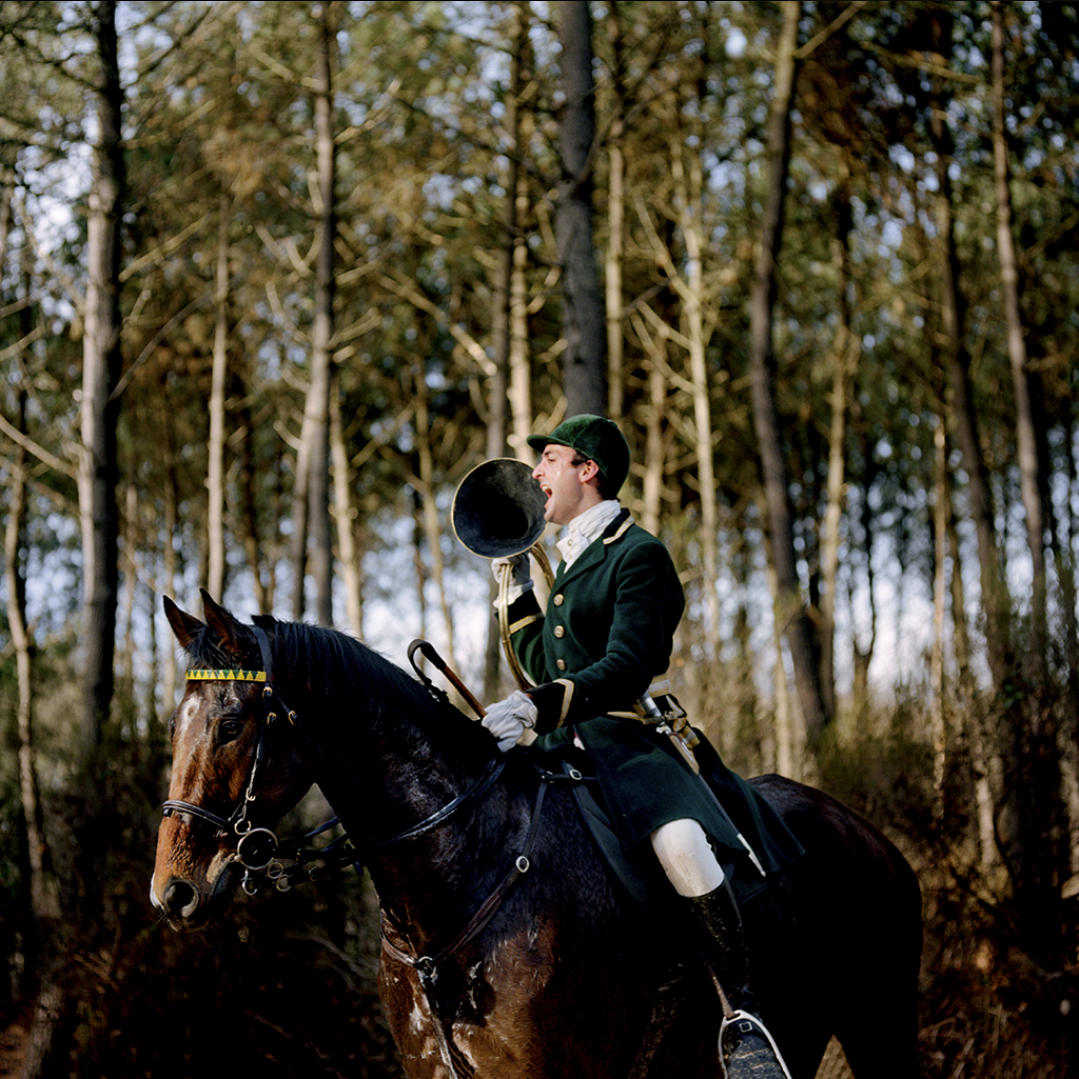 Un homme en uniforme vert et blanc, portant un casque vert, assis à cheval dans une forêt, criant dans un mégaphone