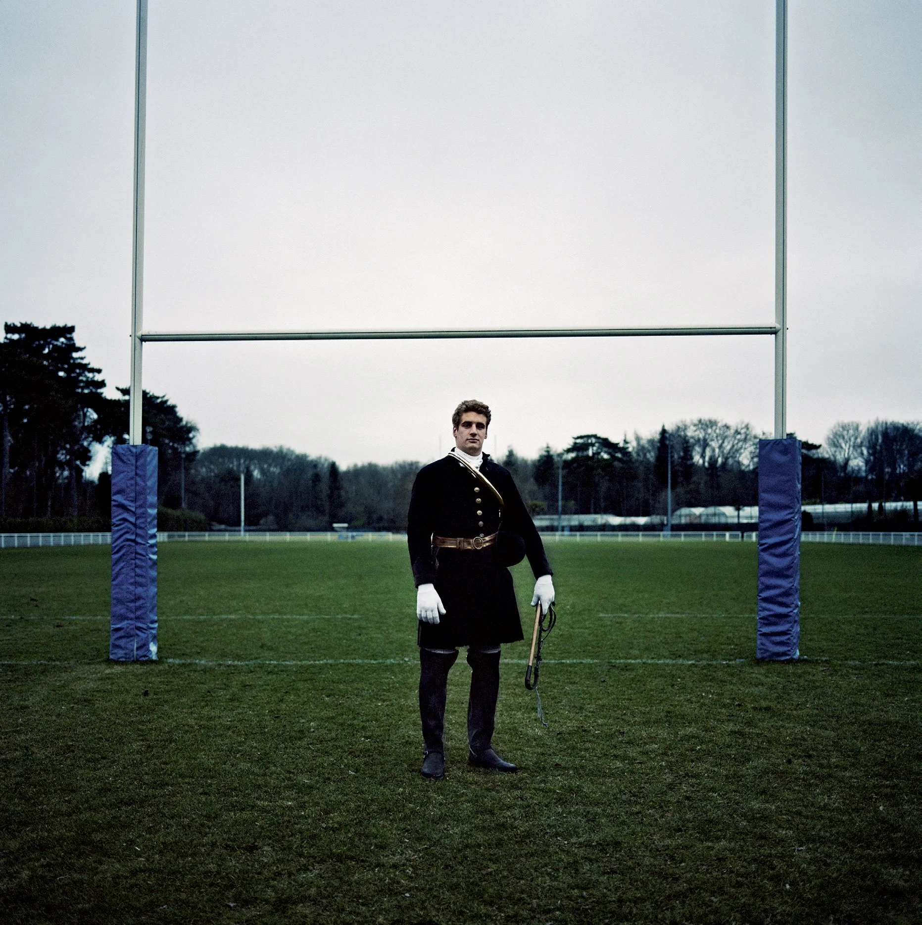 A man in military uniform standing in front of a test post for rugby players on a rugby field"}.