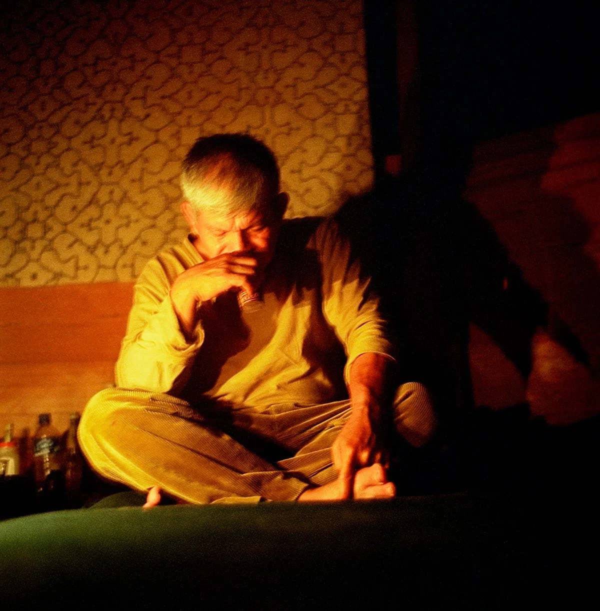 A man in a beige suit sits in front of a decorated wall, holding a cigarette, in a dark, subdued atmosphere.