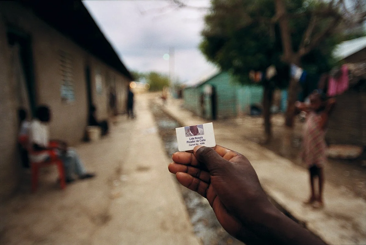 A hand holds an identification card for an anthrax vaccination campaign, in a rural neighborhood with mud houses, people sitting or standing in the street.