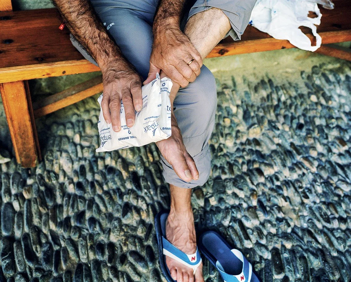 A person sitting on a wooden bench, applying an ice pack to his injured knee, with sandals and a pebbled floor around him.