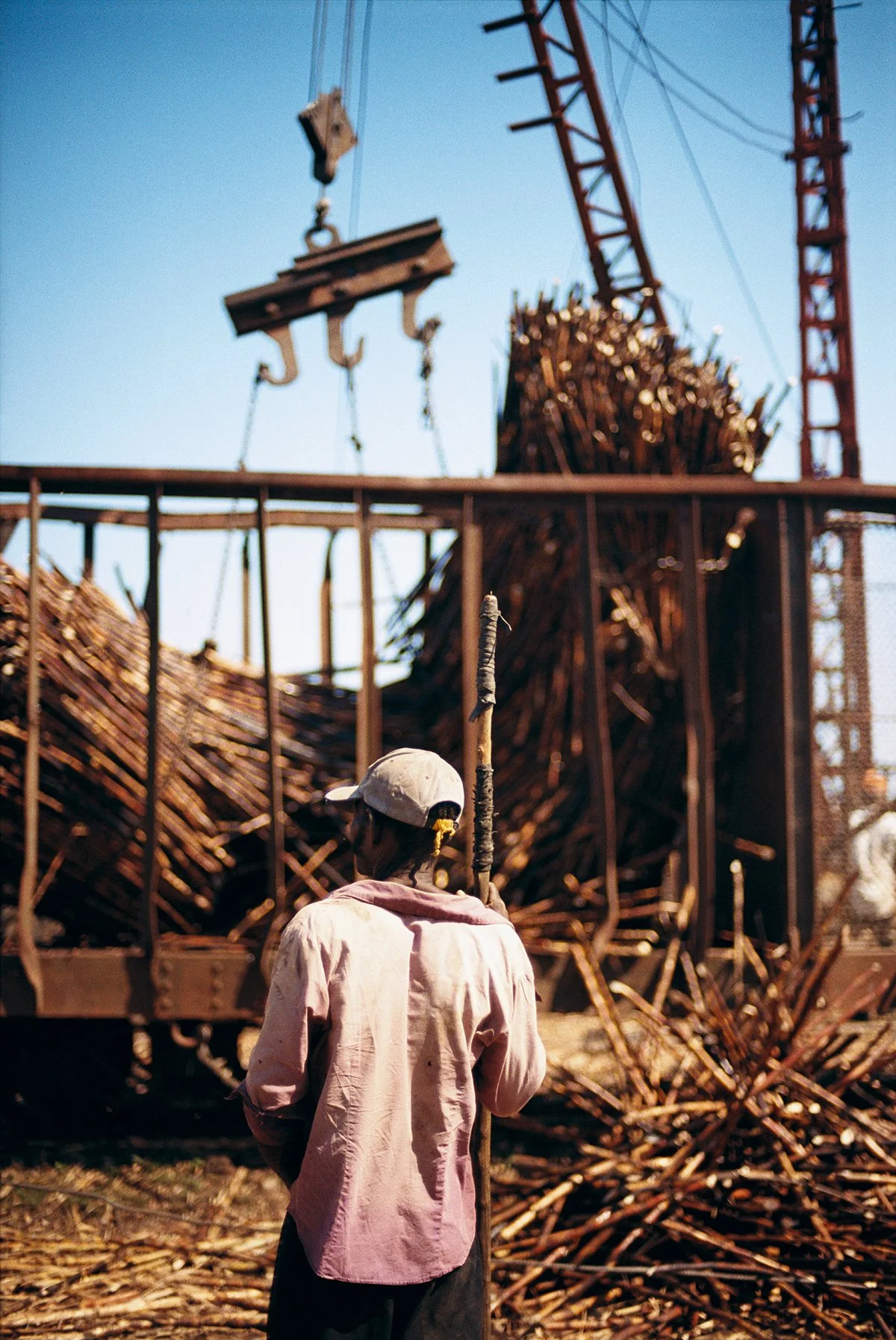 A man in a white hat and pink shirt, from behind, looks up at a large steel structure with lots of stacked pieces of wood or metal, under a clear blue sky.