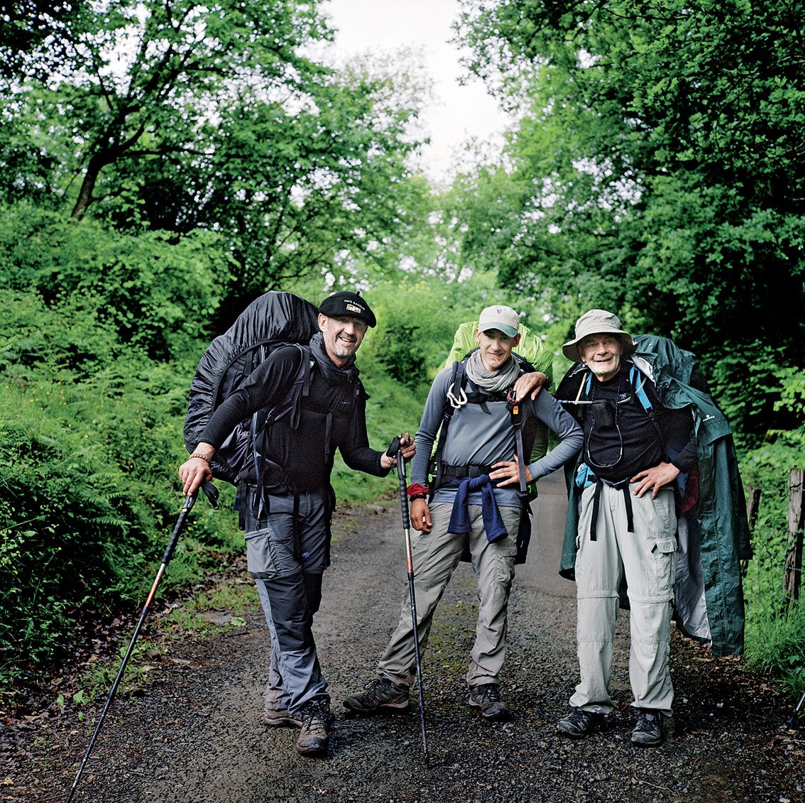Three smiling hikers with backpacks and walking sticks on a green forest trail.