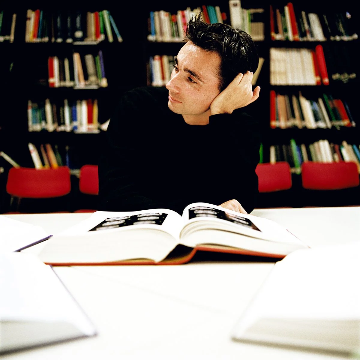 Young man sitting at a table in a library, looking left, with a book open in front of him.