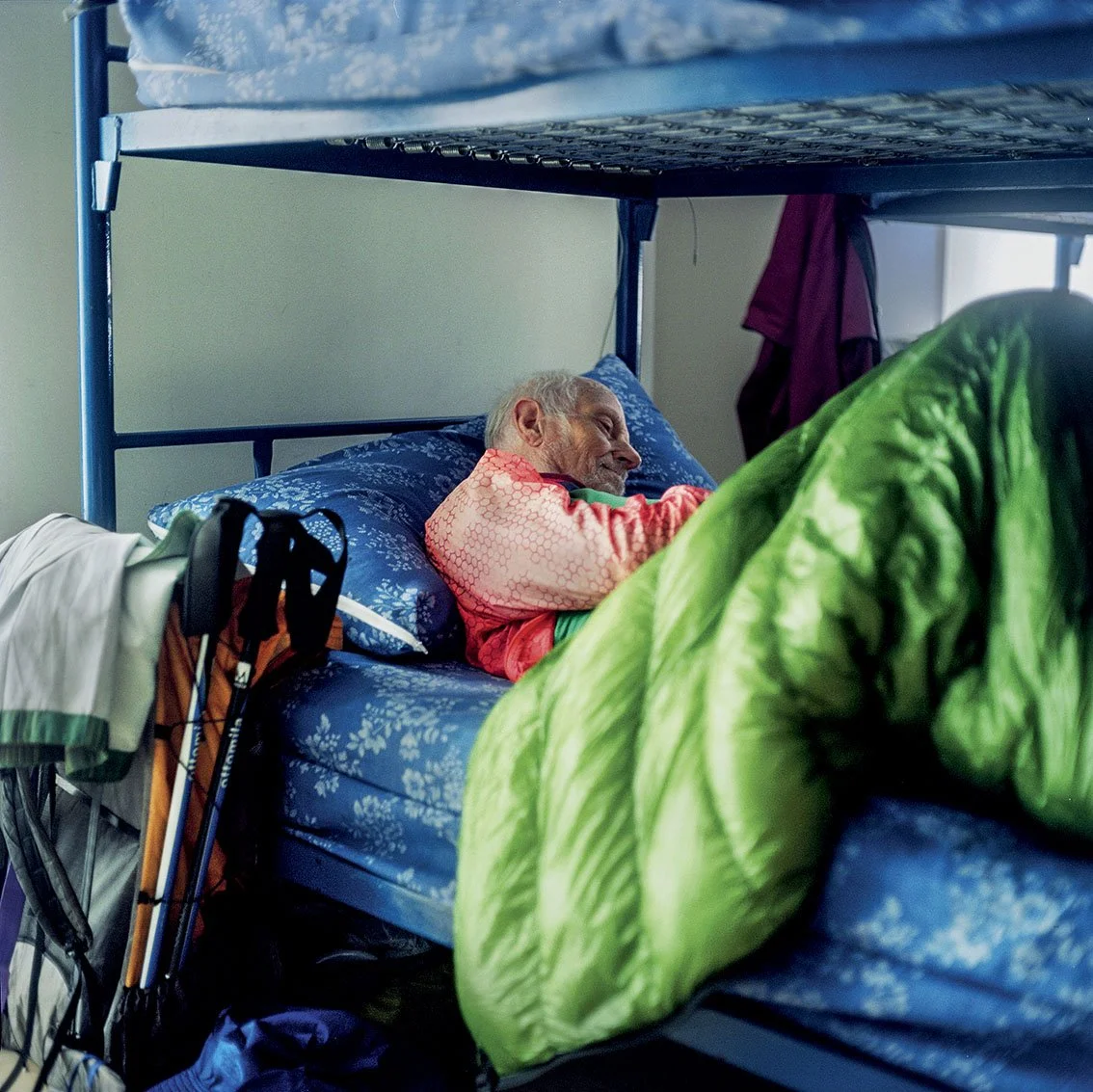 An elderly man sleeping in a bunk bed, wearing an orange sweater, with blue sheets and a patterned green blanket.