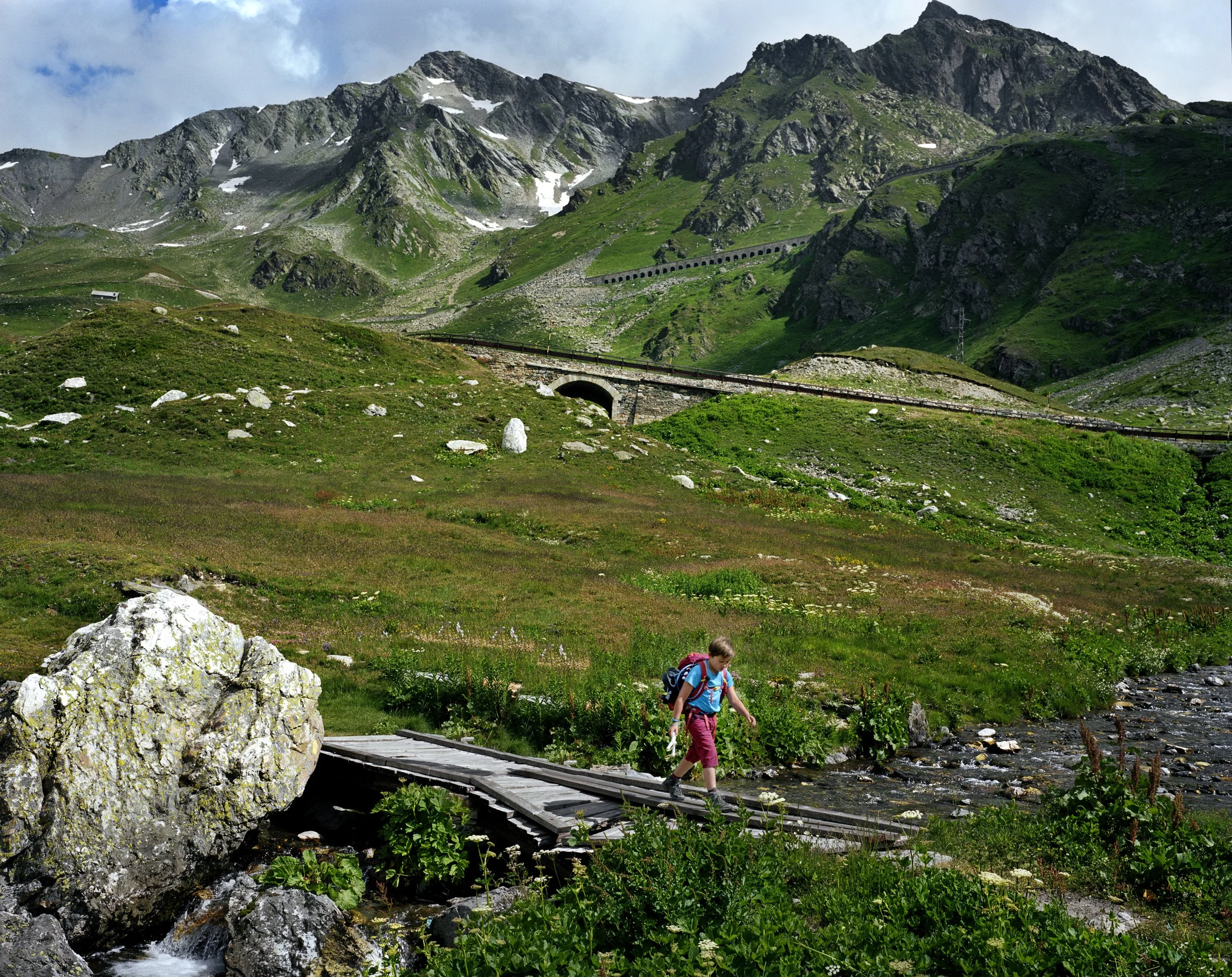 A young girl carrying a backpack crosses a small wooden bridge over a river in a green mountain landscape with snow-capped peaks in the distance.