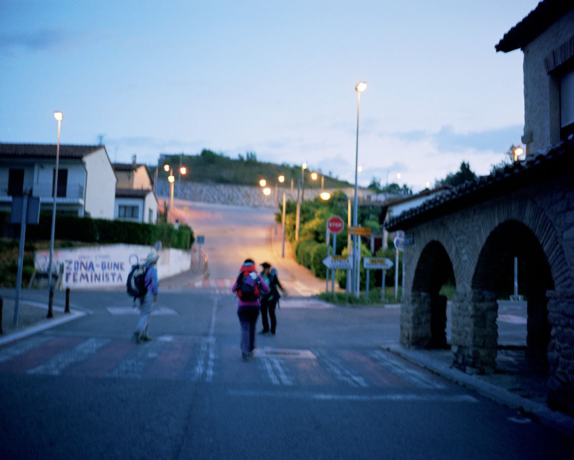 Three people with backpacks crossing an urban crossroads in the evening, stone buildings and streetlamps lit.