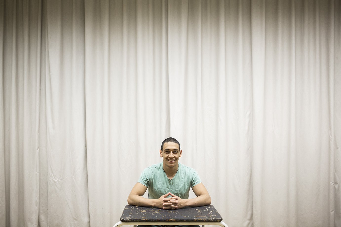 Young man sitting at a table, smiling, in front of a white curtain.
