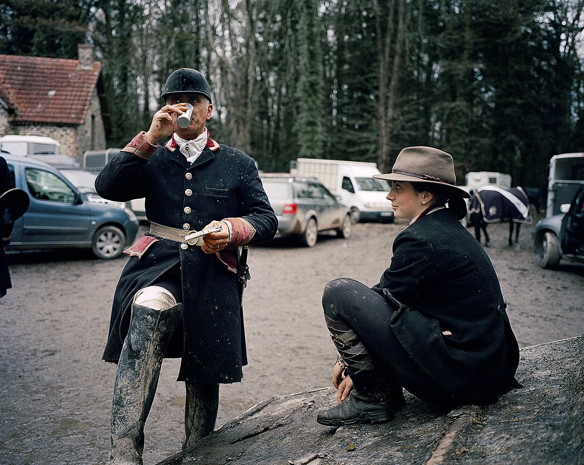 Deux personnes habillées comme des écuyers, l'une en train de boire une boisson et l'autre assise sur un rocher, dans un espace extérieur avec des voitures en arrière-plan.