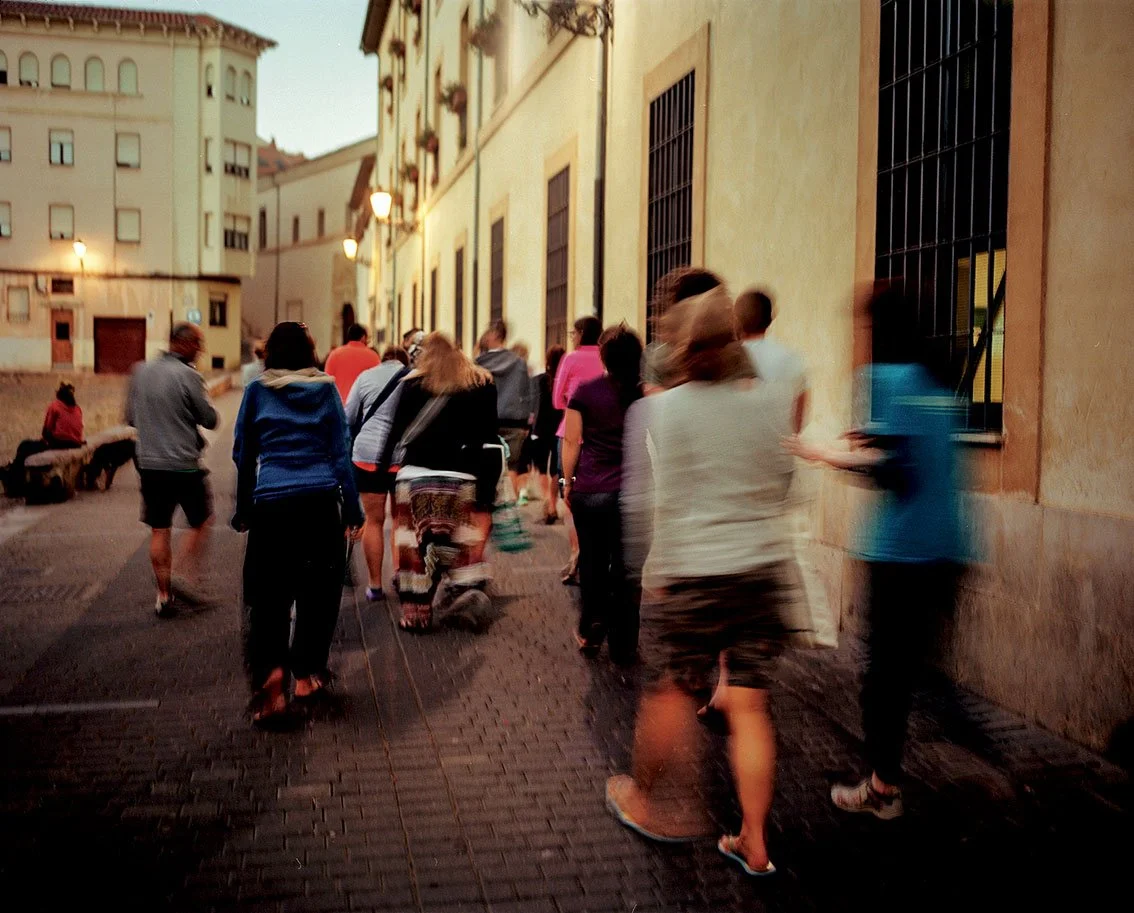 Group of people walking down a cobbled street in the evening, old buildings and street lamps illuminating the scene.