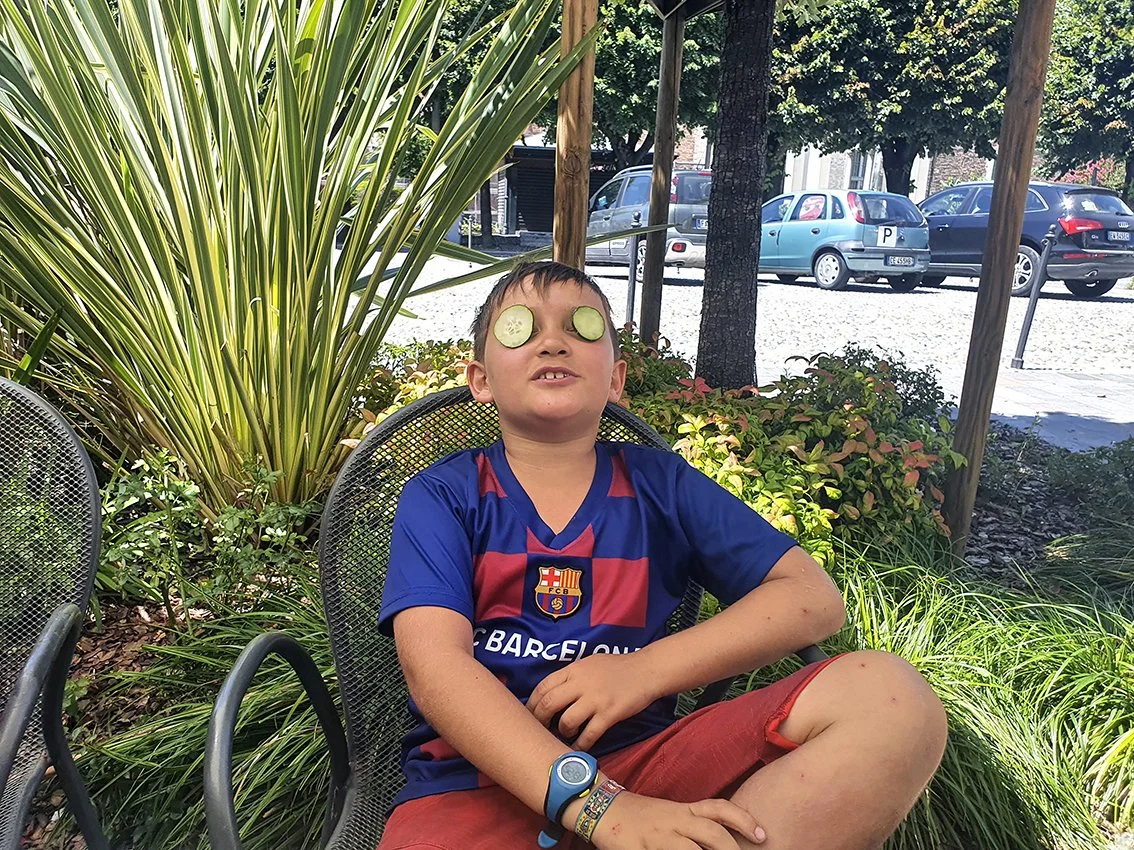 Young boy with cucumber slices over his eyes, sitting outdoors among green plants, wearing an FC Barcelona soccer jersey and a connected bracelet.