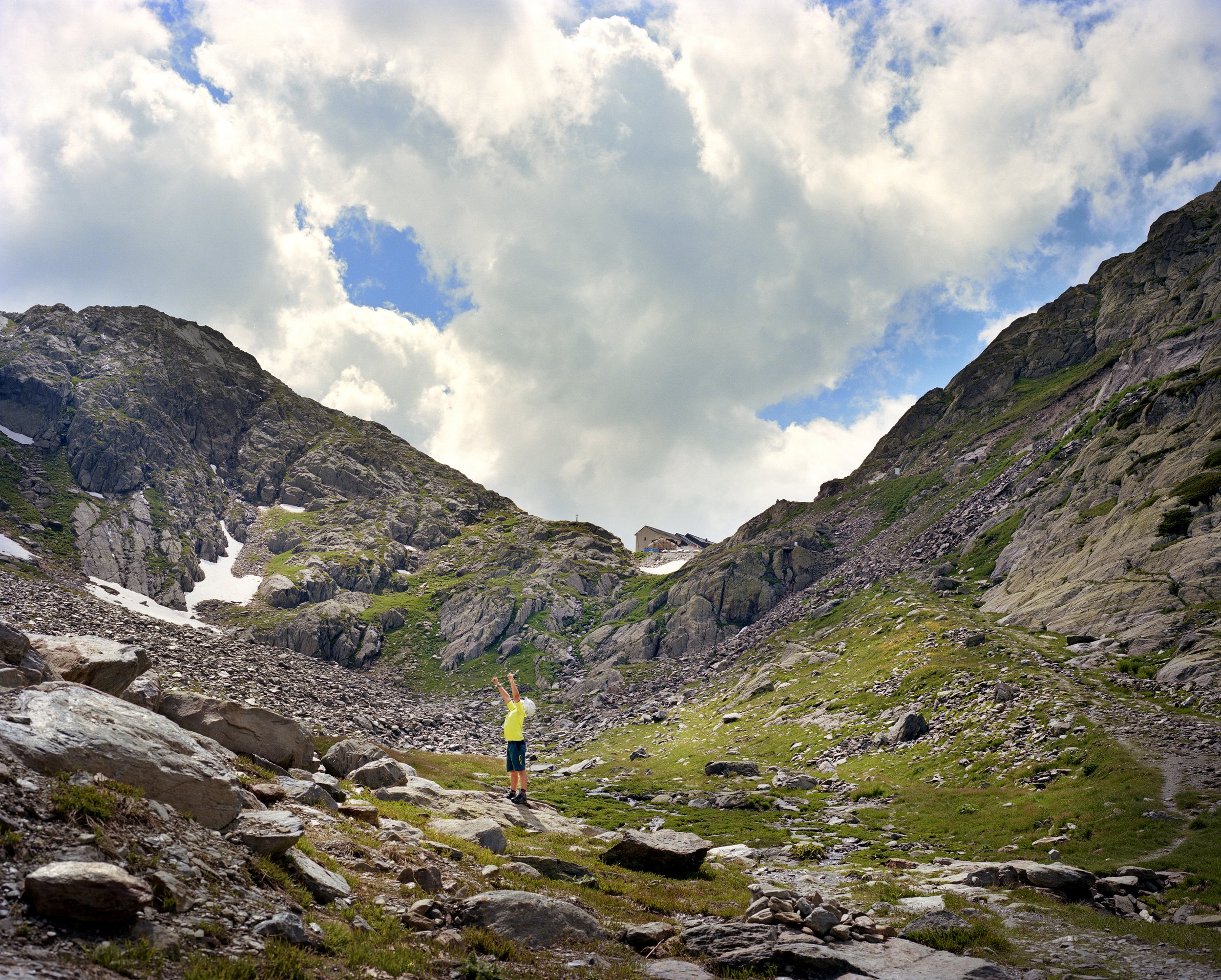 A child in a yellow jacket, in the mountains, under a partly cloudy sky, raising his arms