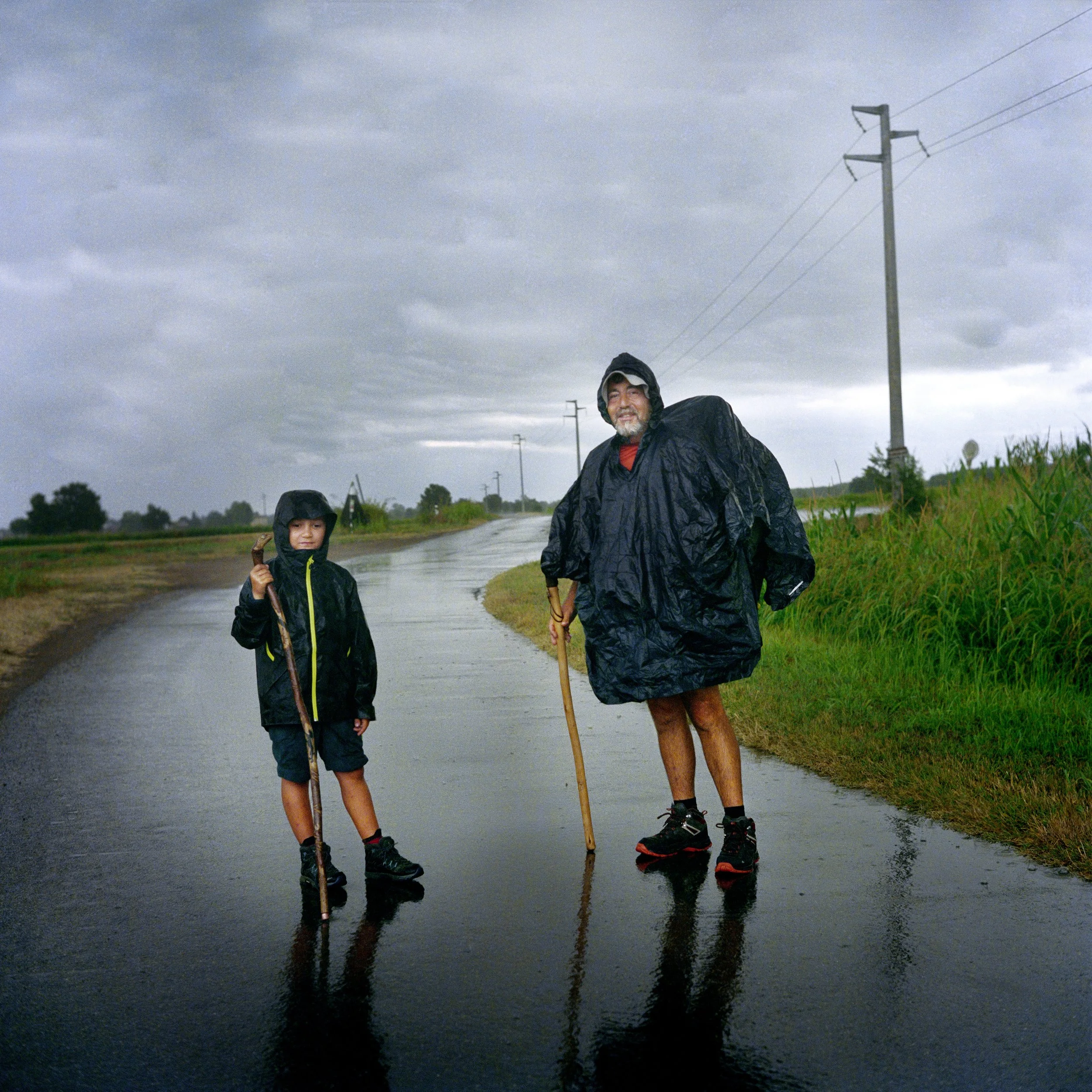 A man and a boy wearing black ponchos stand on a wet road under a stormy sky, holding sticks, surrounded by green fields and power lines.