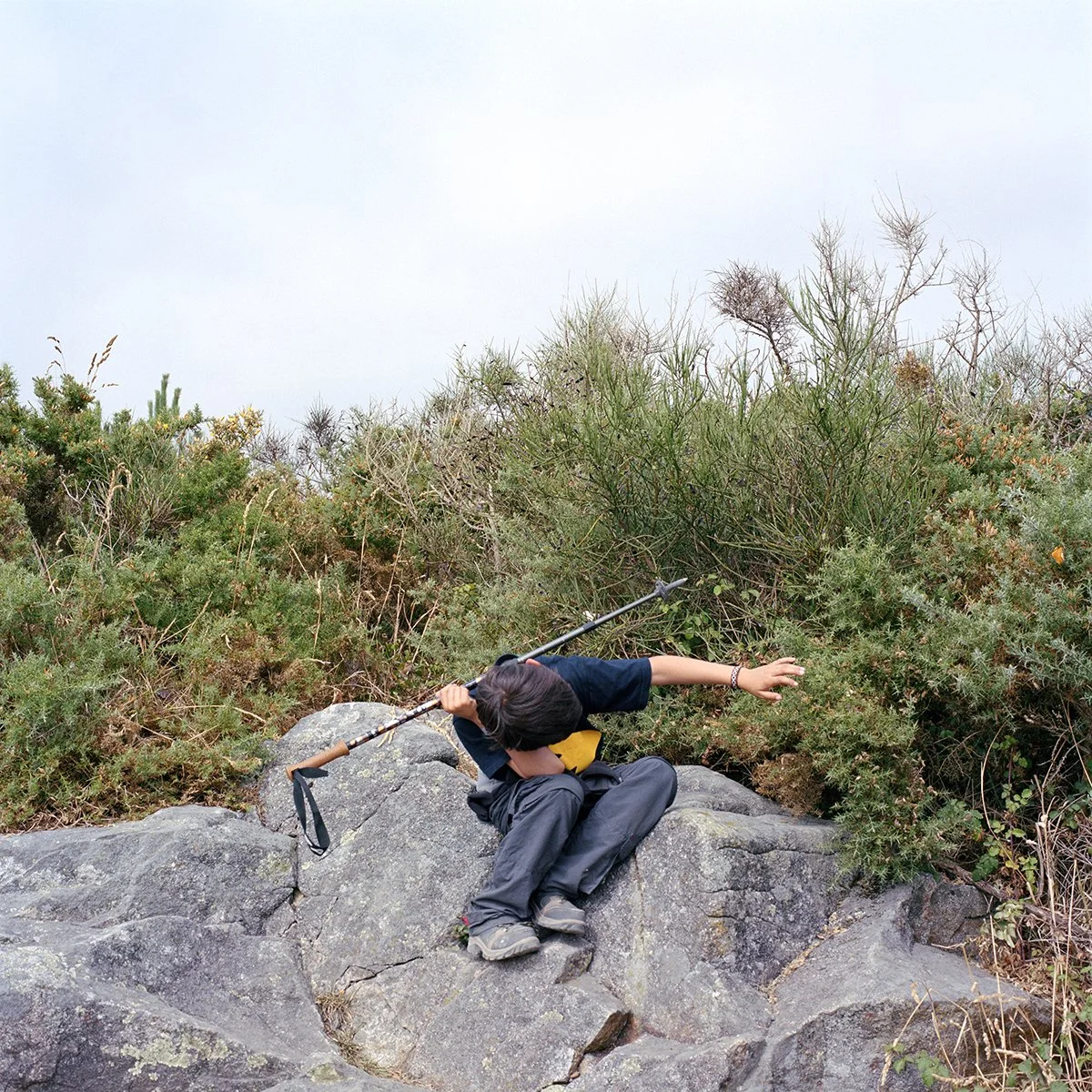 A child sitting on a rock, dressed in black, carrying a stick, in a natural landscape with bushes and a gray sky.