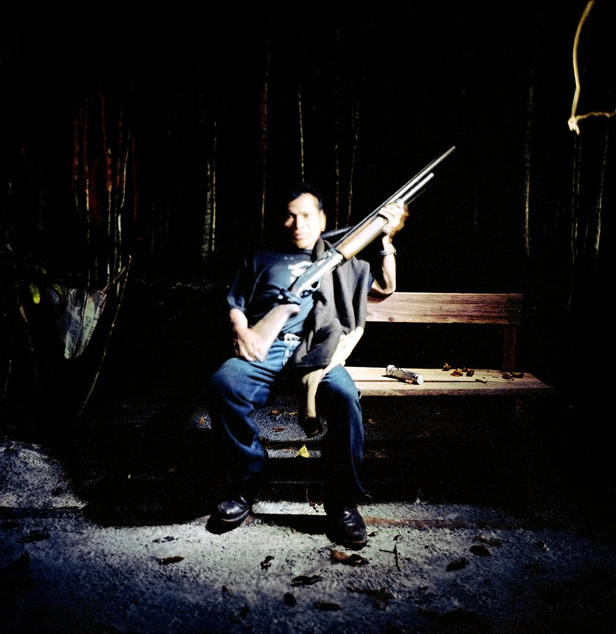 A man sitting on a bench, holding a rifle, in a somber atmosphere. He is wearing a dark jacket and a watch. The scene is dramatically lit, with clothes and objects lying on the bench.