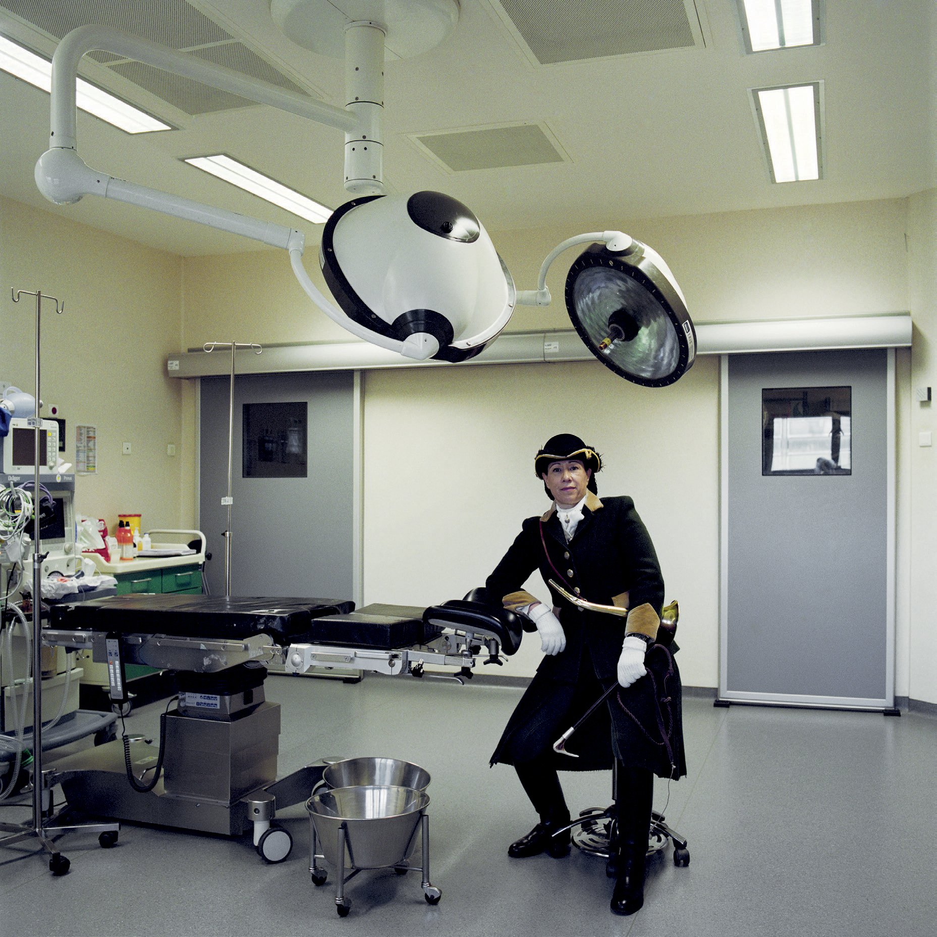 A woman in period dress in a hospital ward, posing with a medical instrument, surrounded by modern medical equipment and surgical lights suspended from the ceiling.