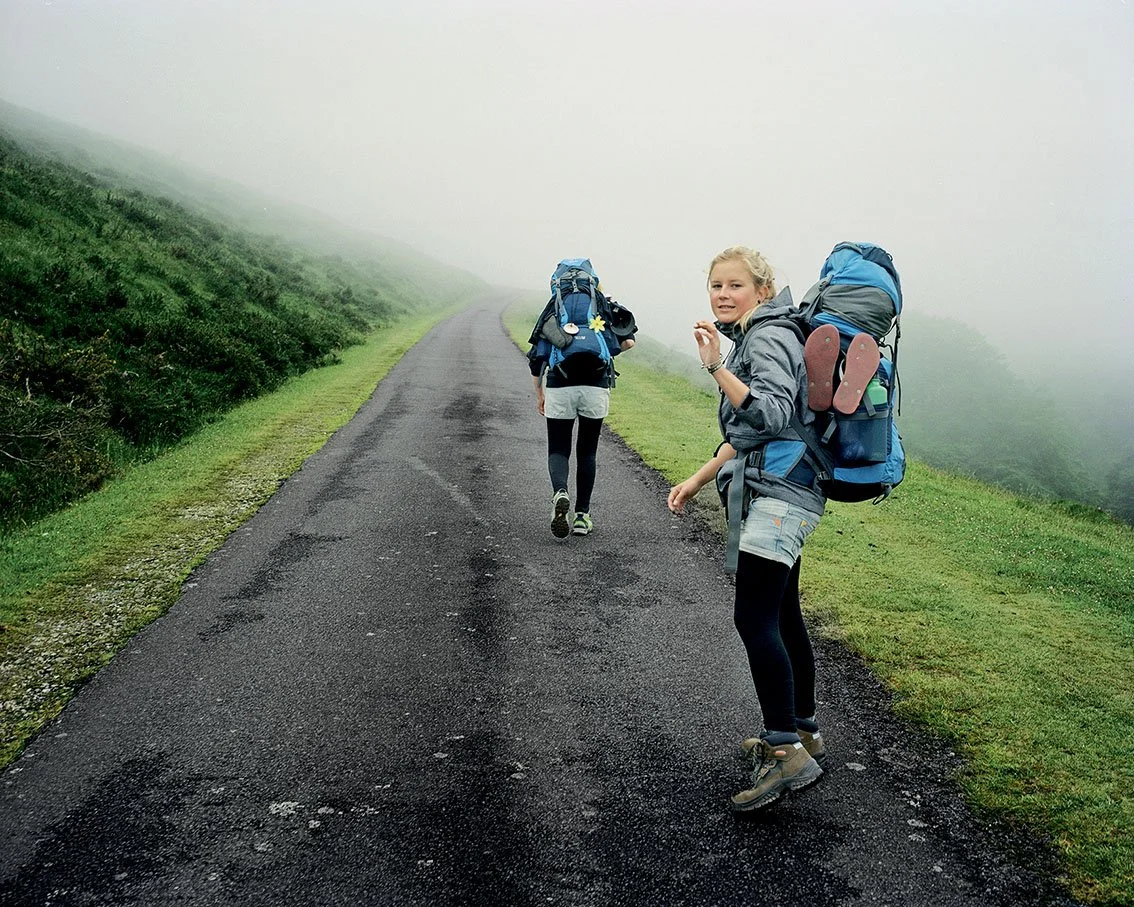 Two young women equipped with backpacks hike a foggy mountain path in trekking clothes.