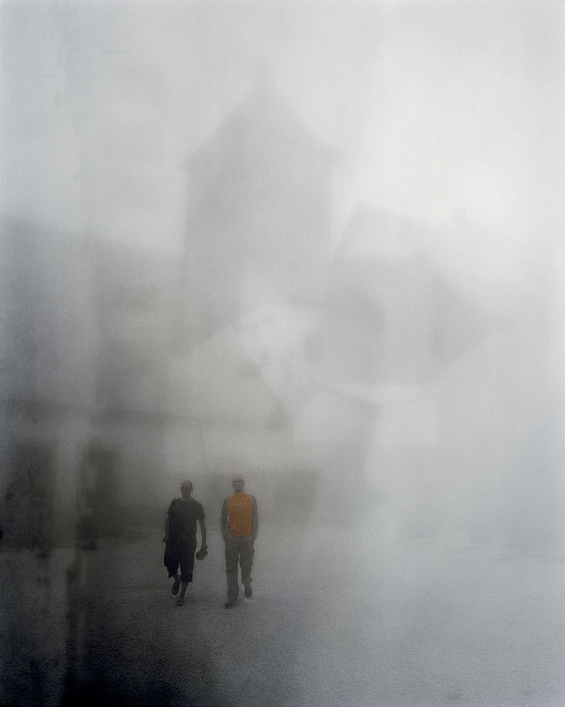 Two people are walking in a foggy or misty area, with a blurred building in the background.