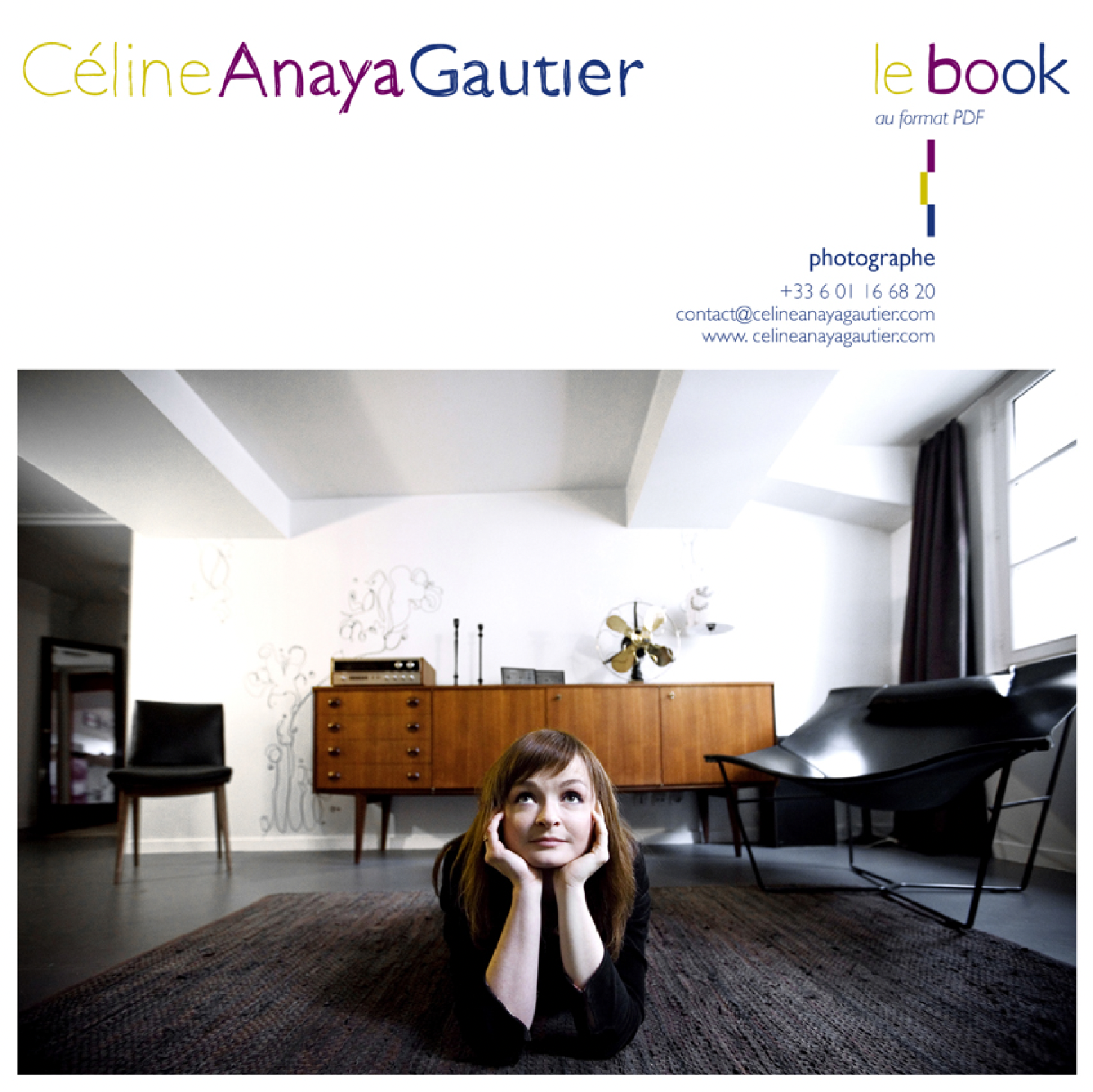 A young woman reclines on a colorful carpet in a living room, looking up. The room is bright, with a large window, wooden furniture, a chair, a floor lamp and minimalist decoration on the wall.