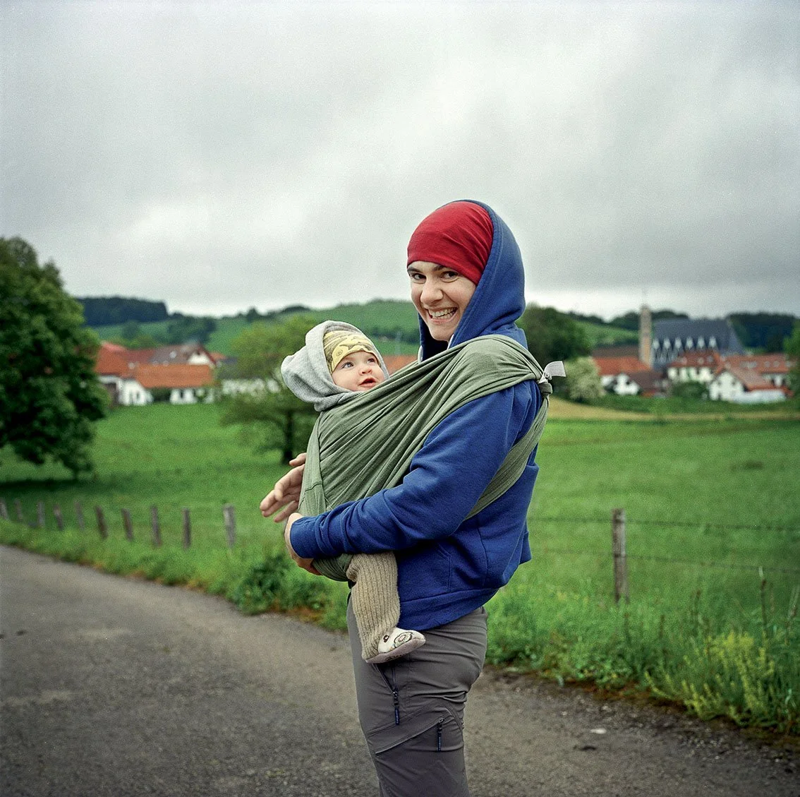 Smiling woman carrying a baby in a sling on a rural road with green fields and houses in the background on a gray day.