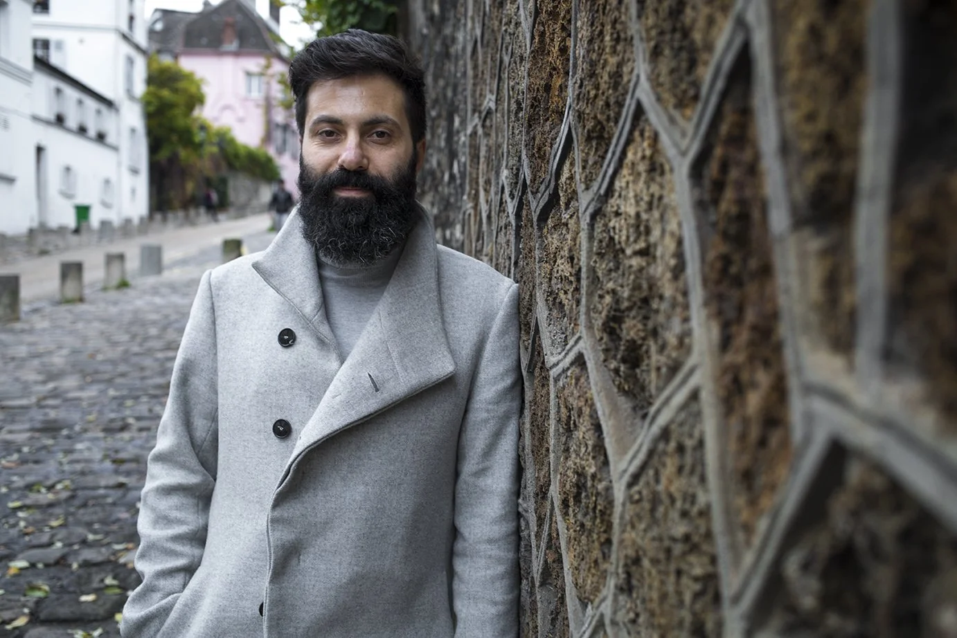 A man with a beard wears a light gray coat and stands next to a decorative stone wall in the open air.