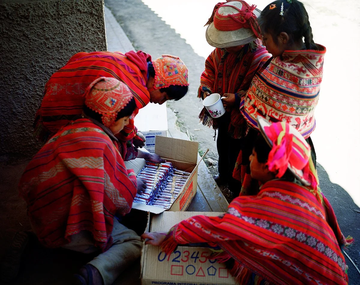 Group of people dressed in colorful traditional clothing, examining objects in a cardboard box. The scene takes place outdoors, near a wall and a water surface.
