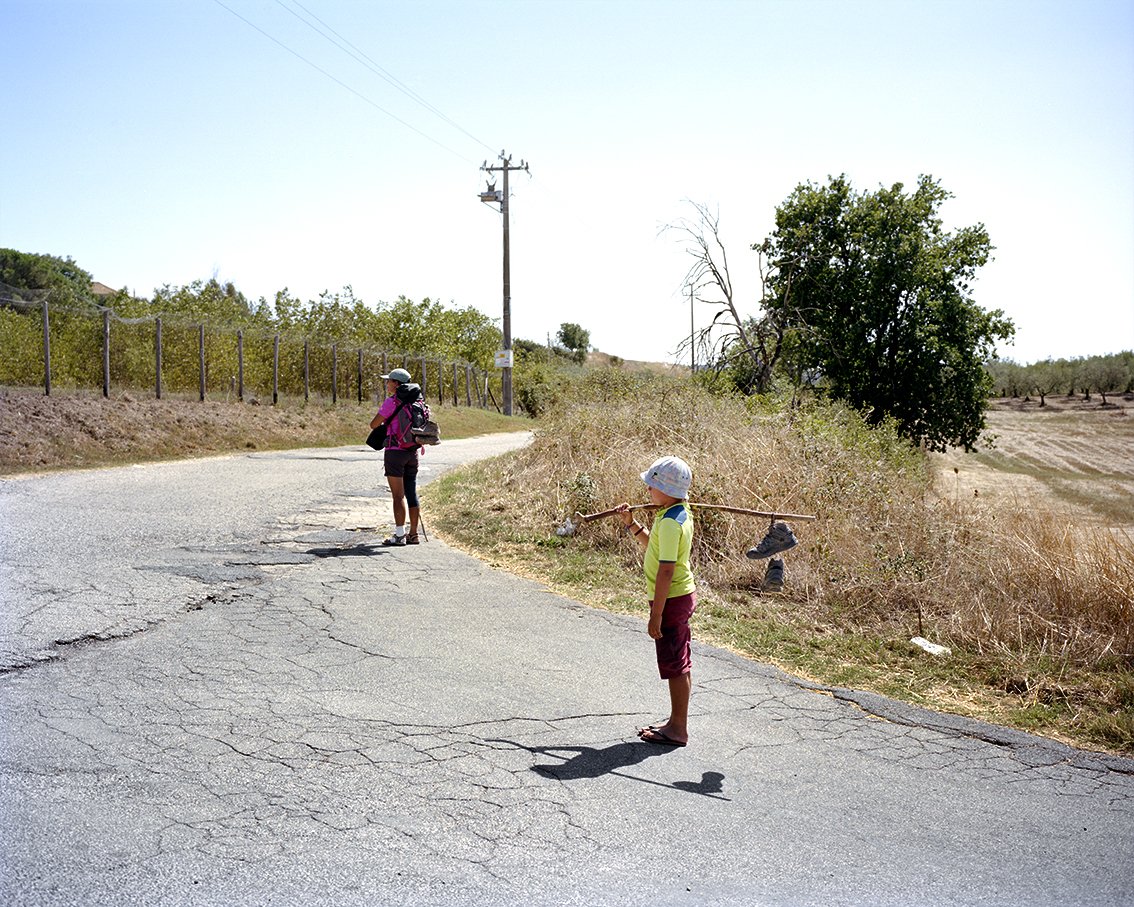 A child in a white hat and green T-shirt, carrying a stick, stands on the cracked road, looking towards a woman with a helmet and backpack on the back, in a hot, dry rural landscape with trees and fields.