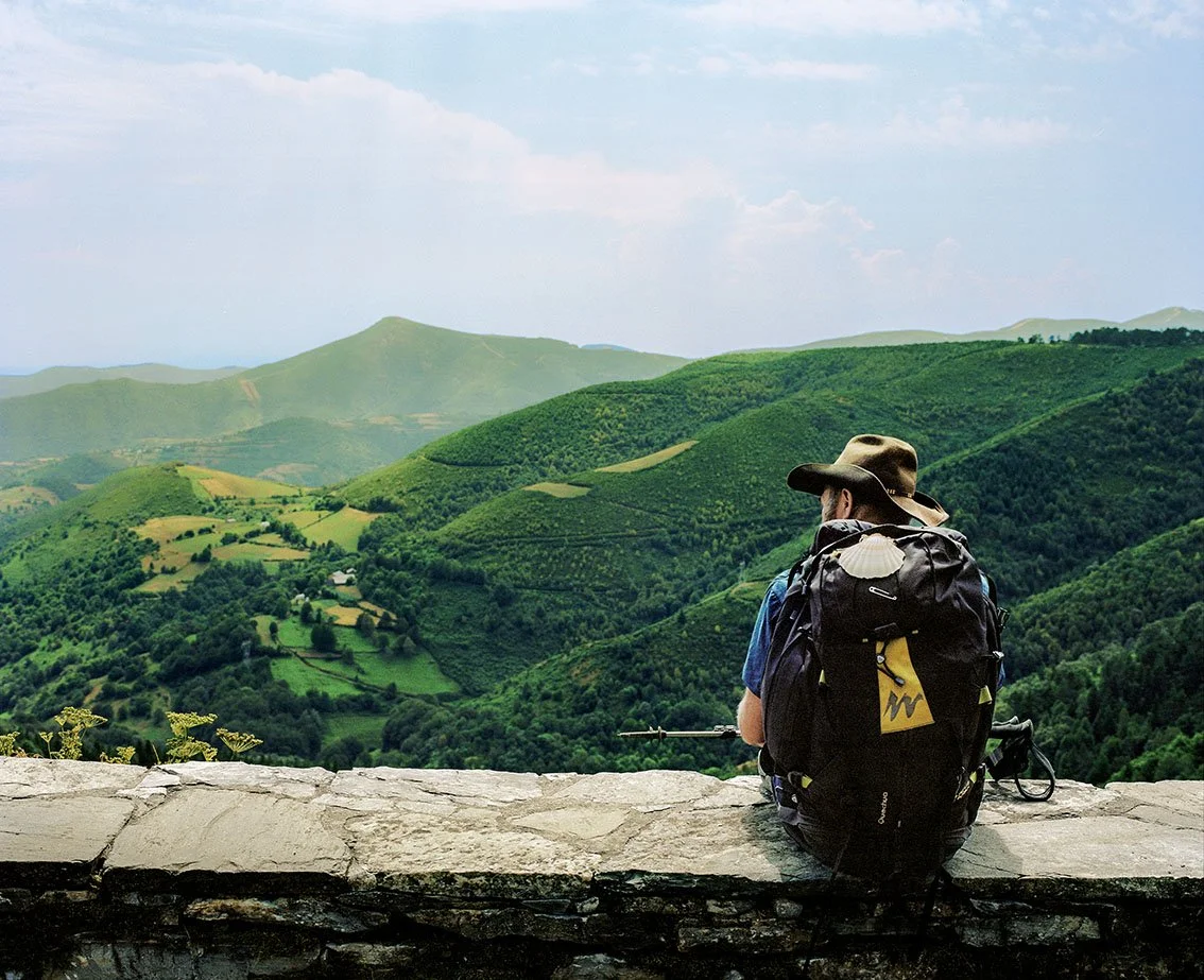 A man with a hat, sitting on a wall, looking at the green mountains in the landscape.