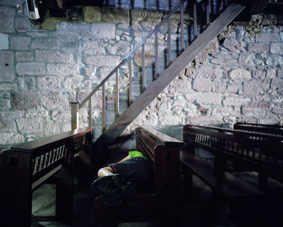 A child in sportswear lying on the floor of a church or old building, looking like a boy sleeping or playing, next to wooden benches, with a stone wall in the background and a wooden staircase to the left.