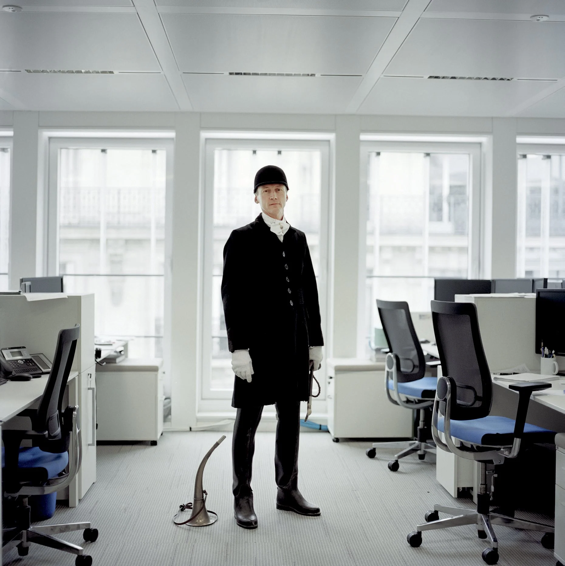 A person dressed as a forest ranger with a black uniform, black hat, white gloves and a pipo, in a modern office with several computers and chairs.