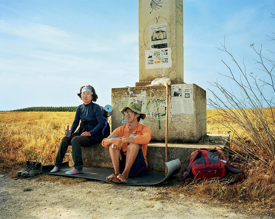 Two hikers sitting in front of a monument in an open field, with backpacks and camping gear.