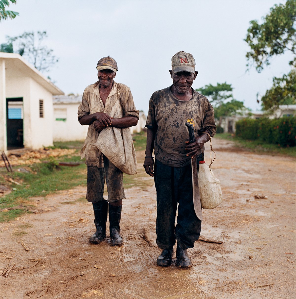 Two elderly men, covered in mud, wearing hats and rubber boots, walking along a dusty path in a rural village, one of them holding a tool.