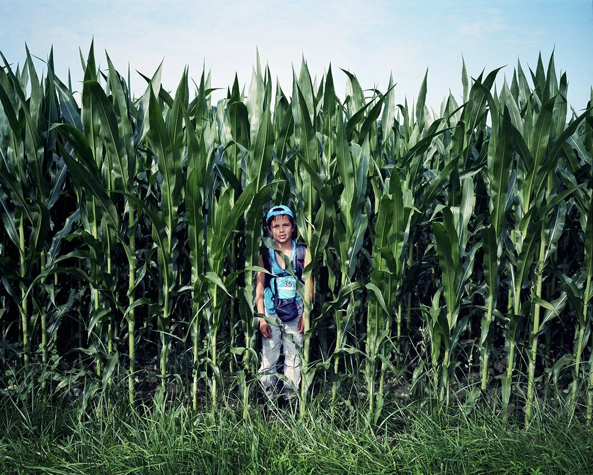A young boy in a hat, carrying a backpack, stands in the middle of a green cornfield.