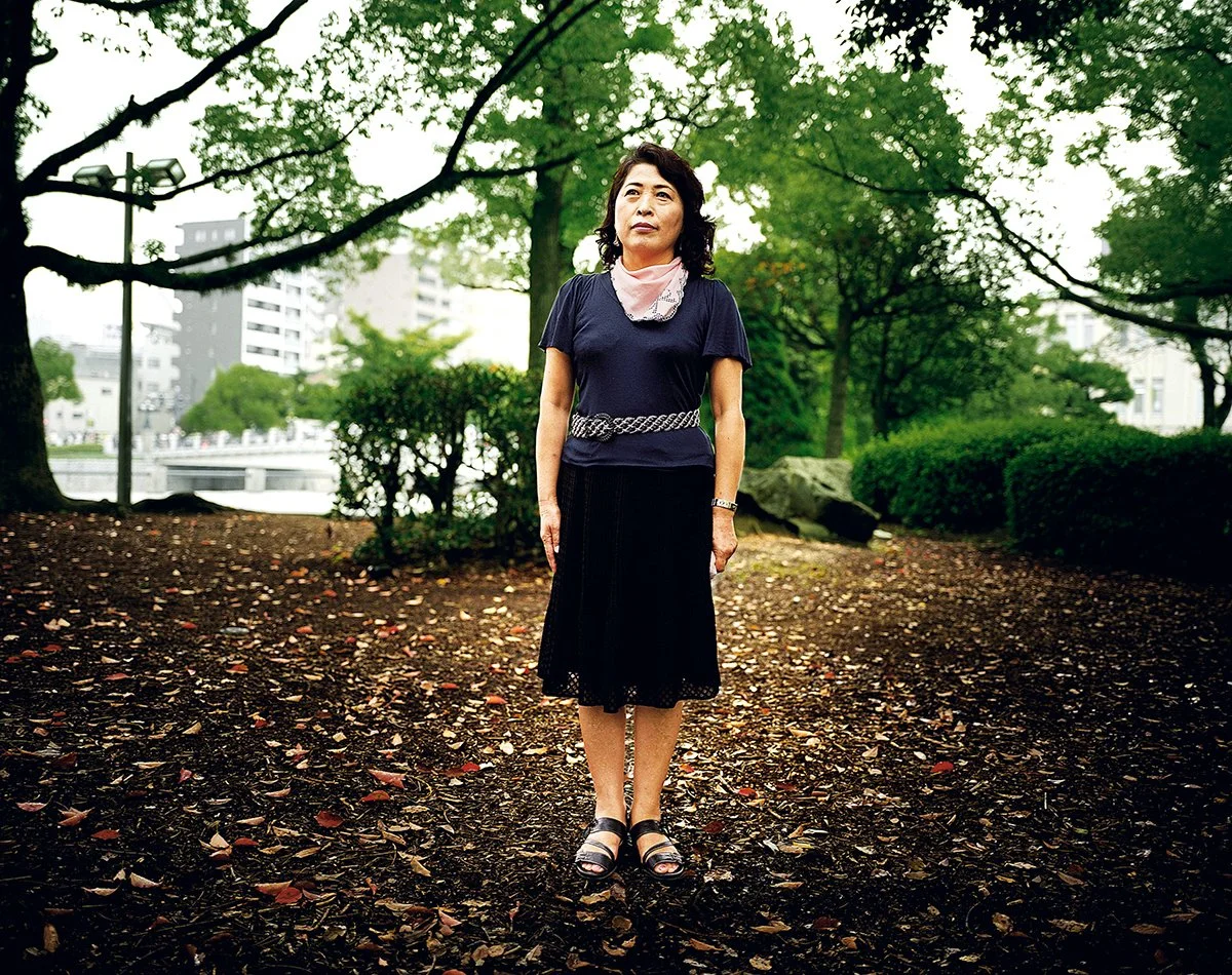 Une femme debout dans un parc urbain avec des arbres verts et des bâtiments en arrière-plan.