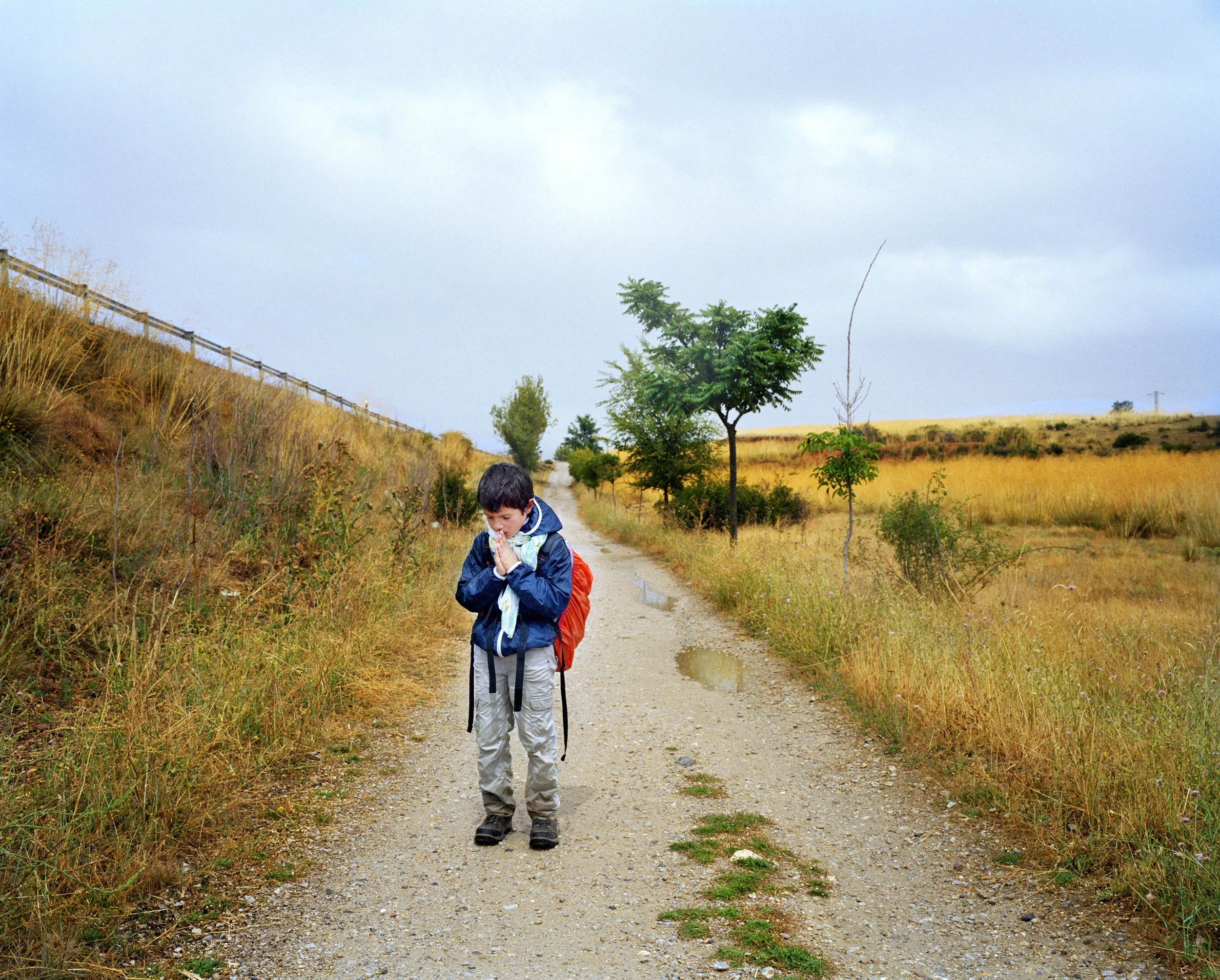 Un garçon marche seul sur un chemin de terre dans un paysage rural avec des arbres, de l'herbe sèche et un ciel nuageux. Il porte un sac à dos orange et une veste bleue, et il a la tête baissée avec les mains jointes.
