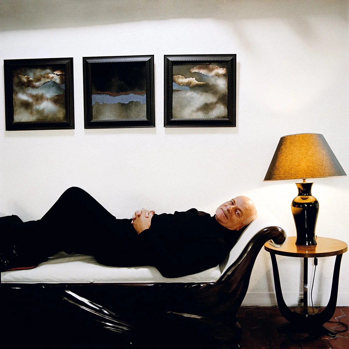 A man lying on a bed in a room with cloud paintings on the wall, a table lamp lit beside him.