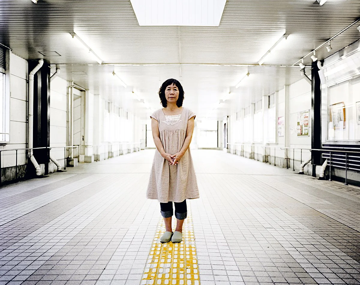 Une femme se tenant au centre d'un passage couvert intérieur, portant une robe beige, des jeans roulés et des chaussures gris clair, dans un environnement lumineux.