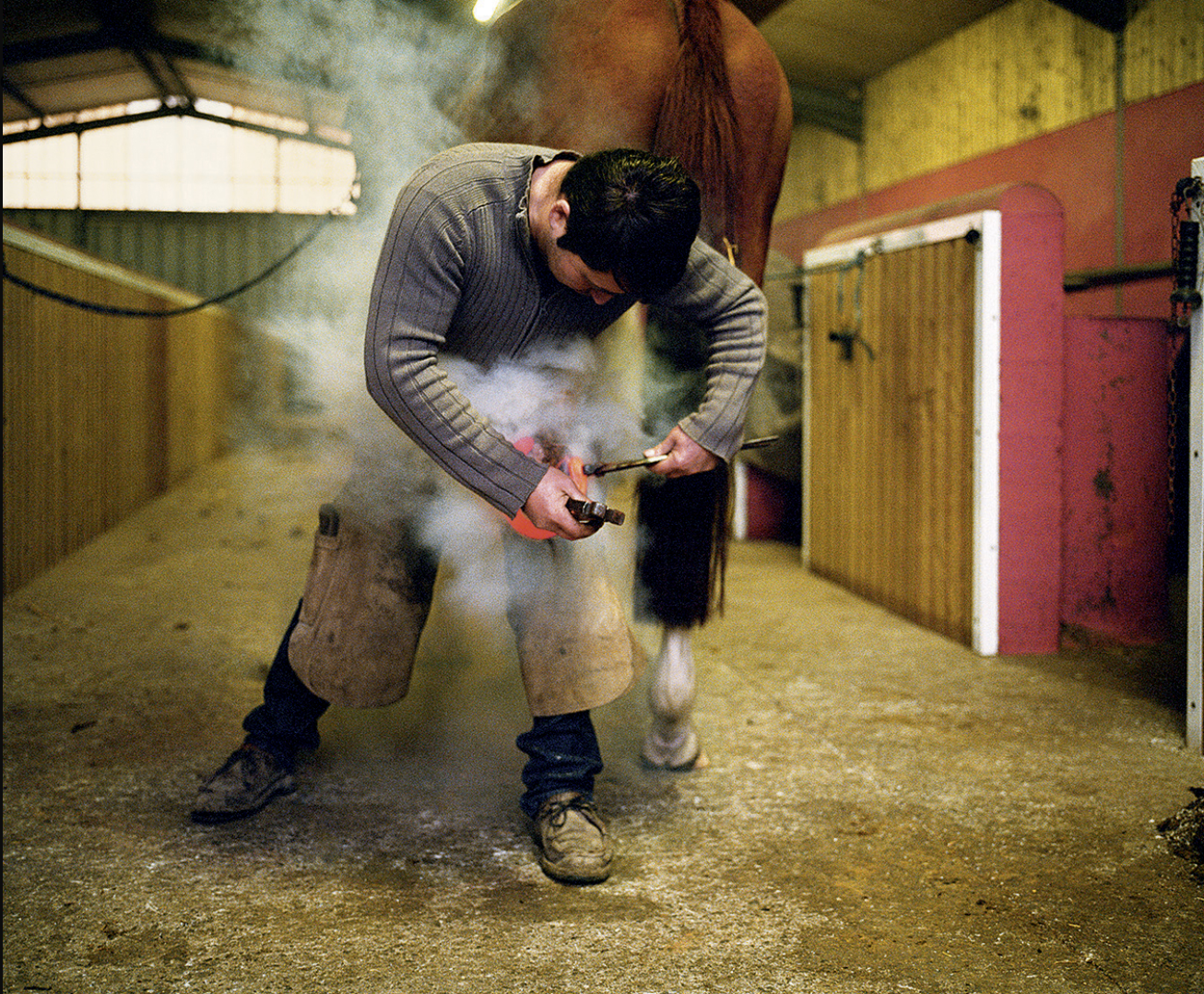 Un homme en train de forger dans un atelier avec de la fumée dense, une étable en arrière-plan.