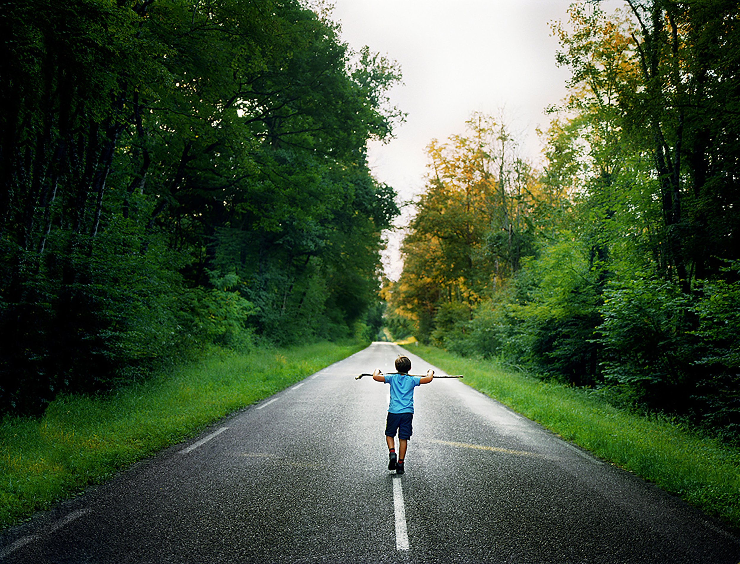 Un enfant court seul sur une route bordée d'arbres verts, mais le fond est brumeux et gris, suggérant une pluie ou un brouillard léger.