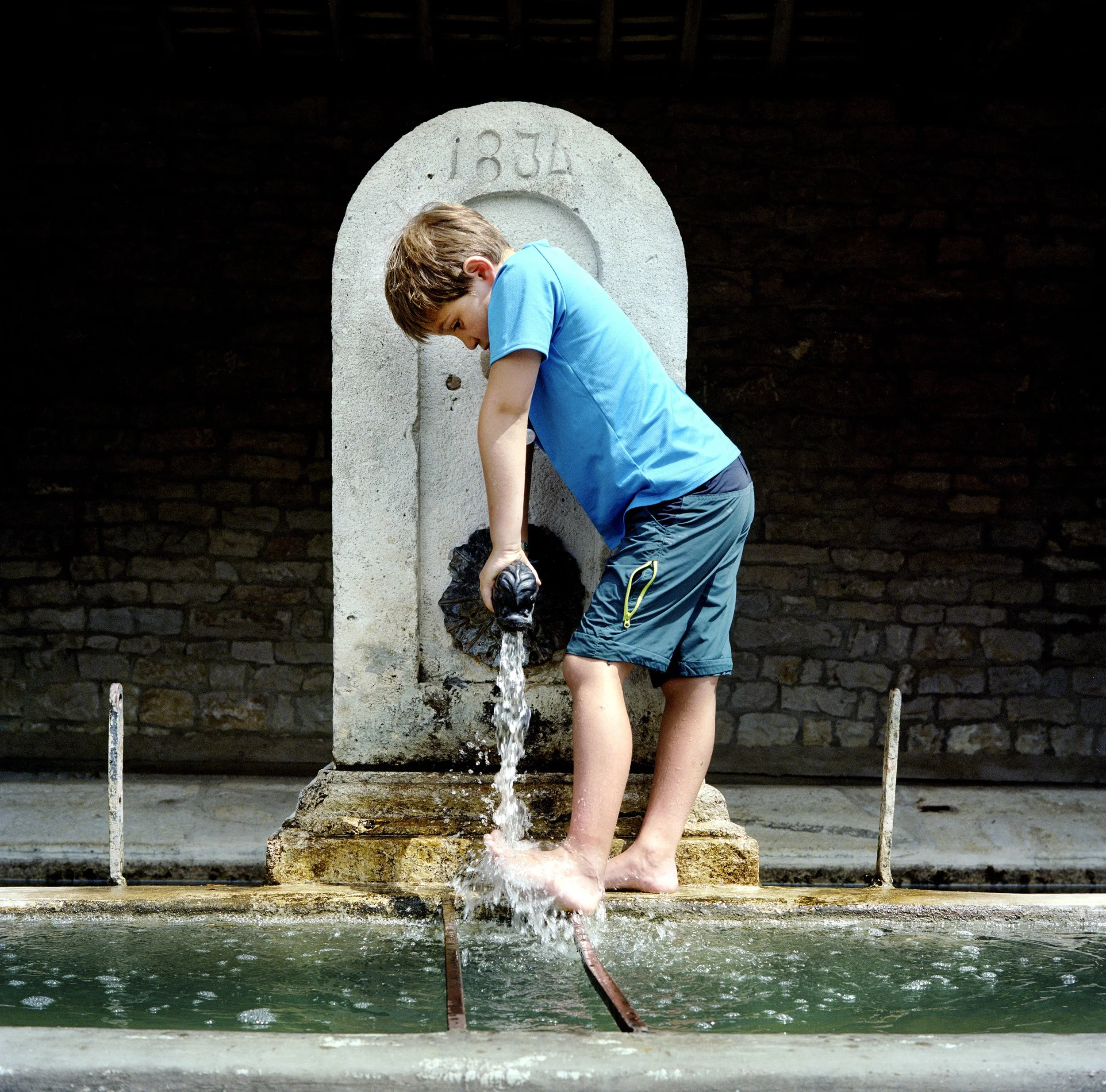 Un garçon brun, portant un t-shirt bleu et un short côtelé, est en train de remplir une bassine d'eau sous une fontaine en pierre avec une plaque portant la date 1834, dans un environnement à l'abris, probablement un musée ou une vieille ville.