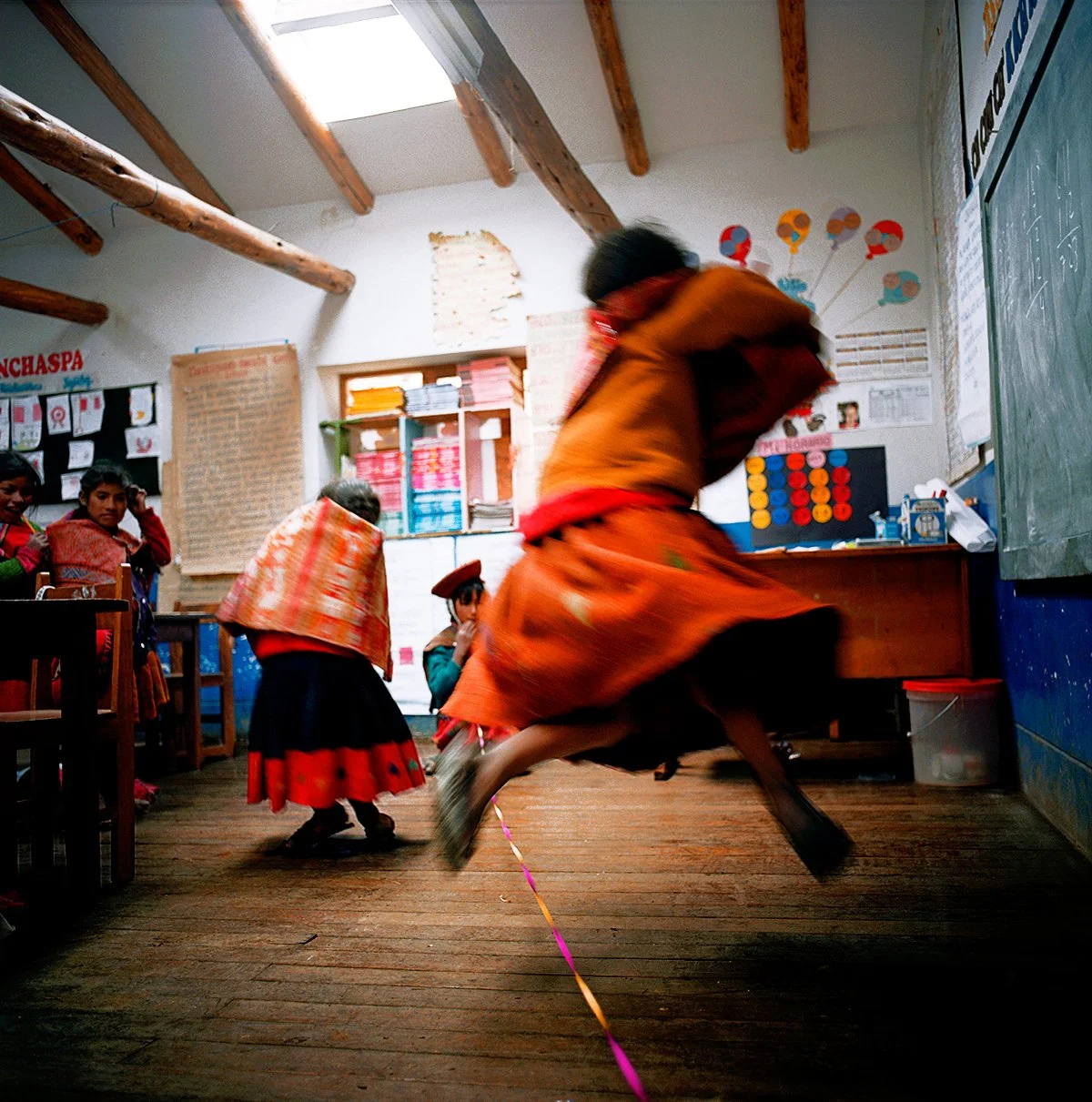 Children in colorful traditional dress play in a classroom with pictures and wall decorations, including a moving person skipping a skipping rope.