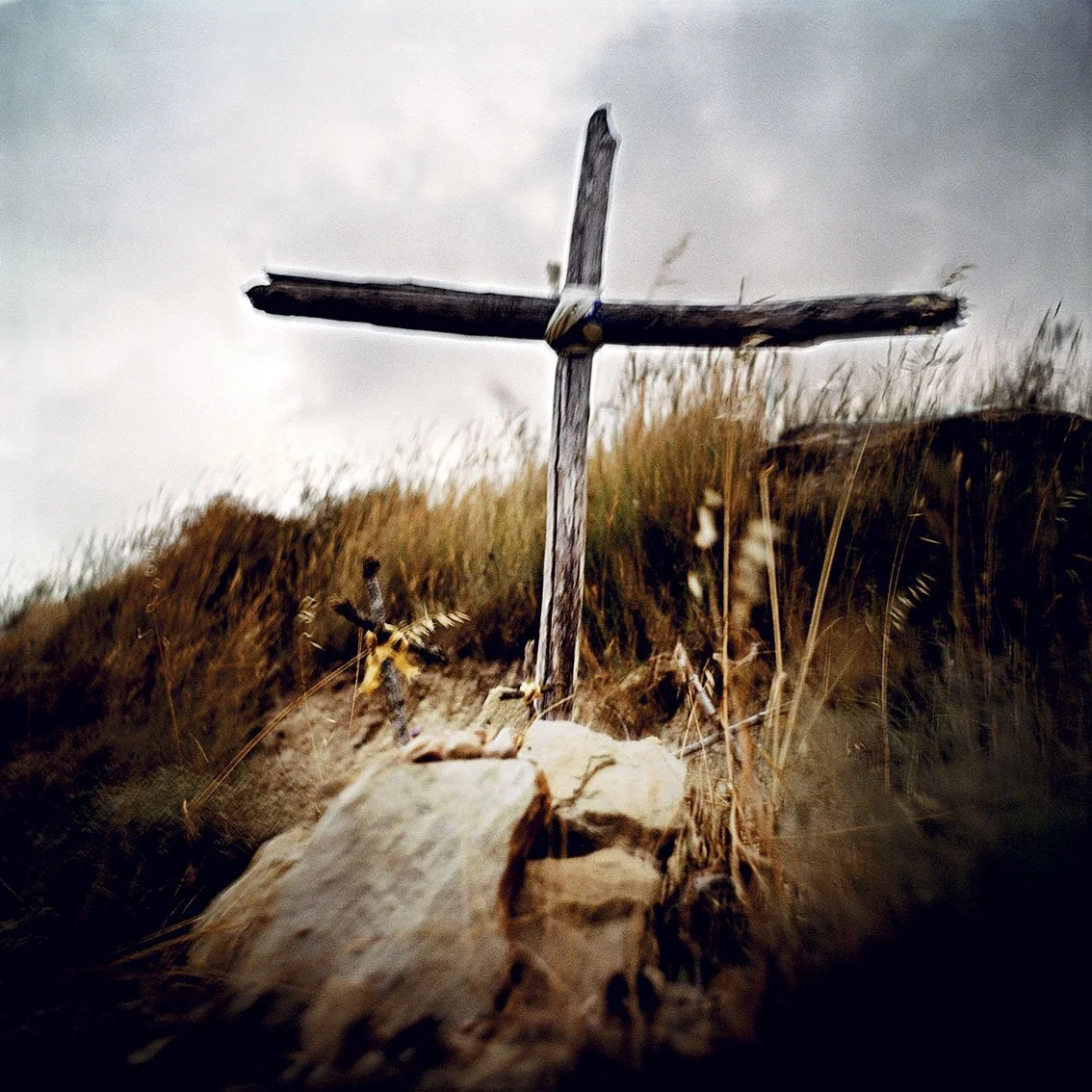 Rudimentary wooden cross planted in rocky terrain with dry grass, under a cloudy sky.