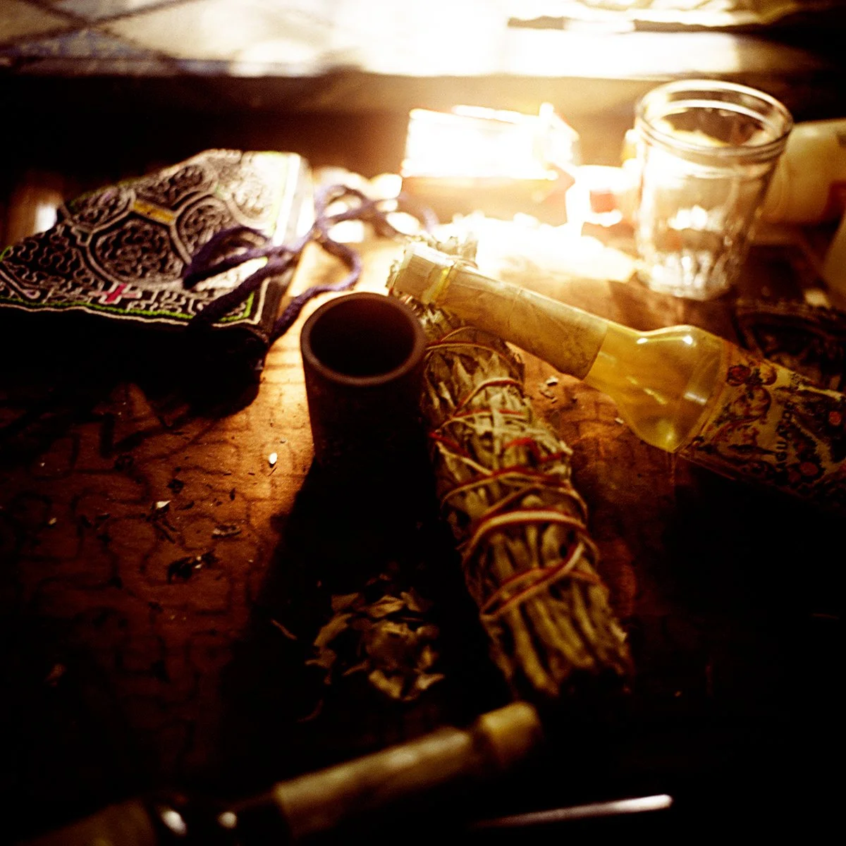 Witchcraft objects on a wooden table, including a wand, herbs, a bottle of oil, a glass, and a patterned cloth, under a warm light.