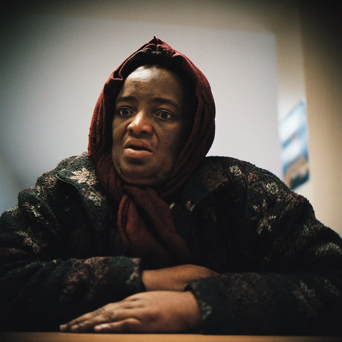 Une femme semble préoccupée ou inquiète, assise à une table, portant une tête enroulée dans un foulard rouge et un manteau noir.