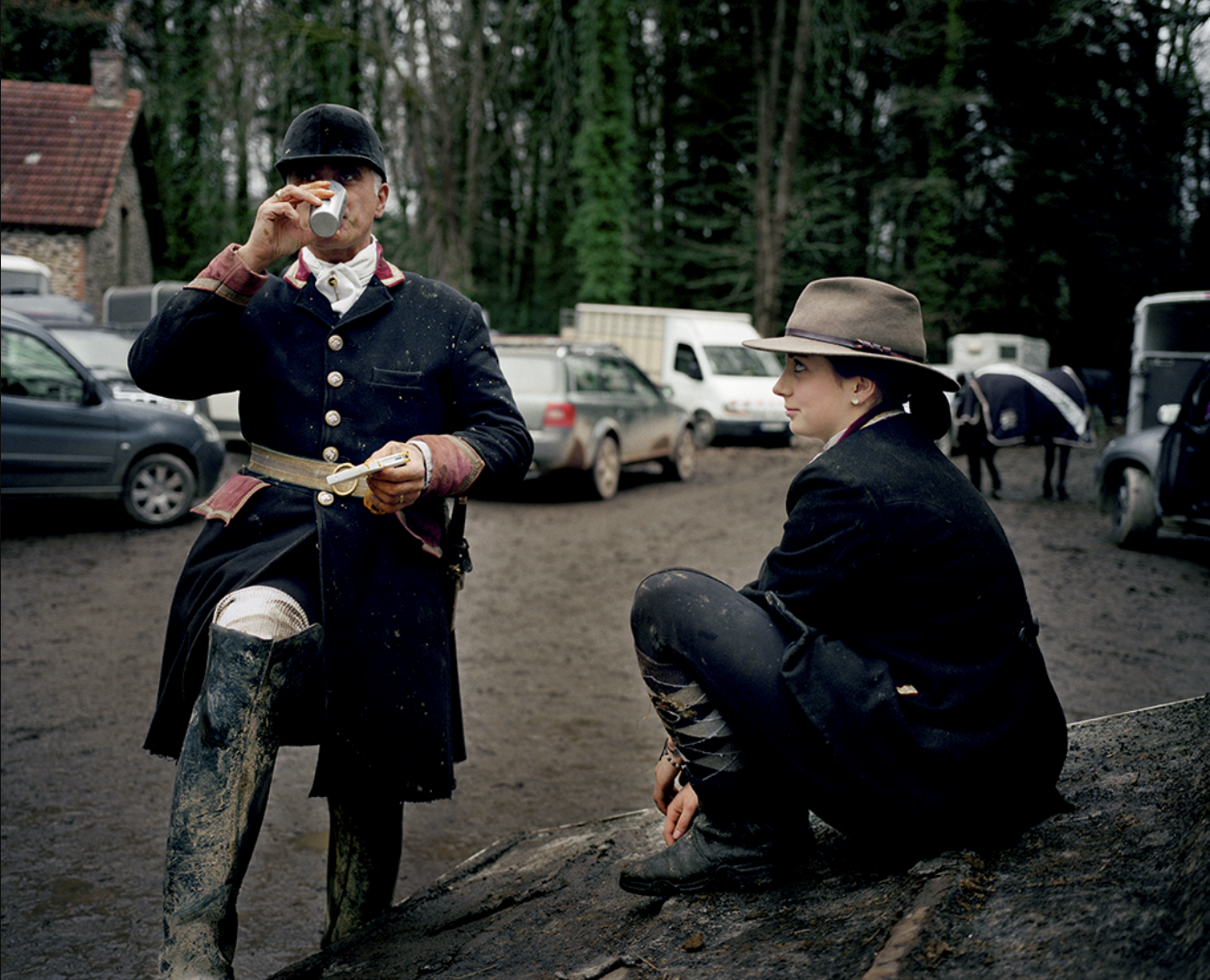 Deux personnes en costume d'époque dans un parking forestier, une homme buvant dans une boîte de conserve, une femme assise sur un rocher, entourés de voitures et de cavaliers.
