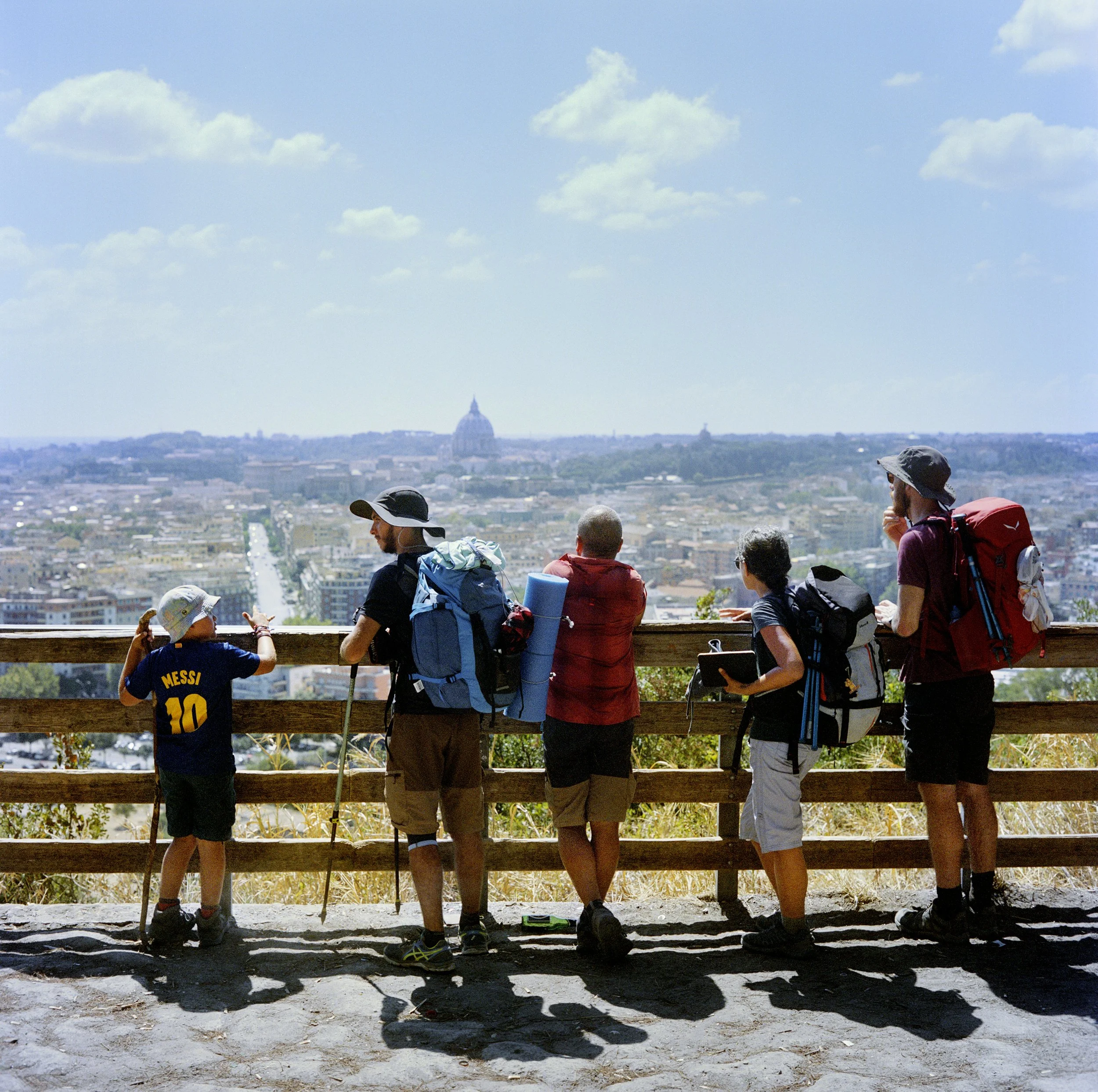 A group of five backpackers looking out over the panoramic view of Paris, with the Sacré-Coeur visible in the distance, on a sunny day.
