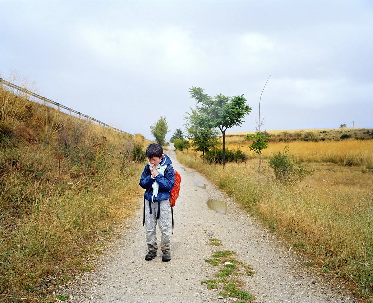 A boy walks alone along a dirt road in a rural landscape with grasses and a few trees, on a cloudy day.
