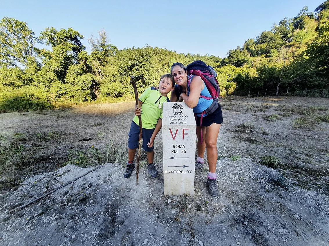 A woman and a young boy pose next to a signpost during a nature hike. The woman is wearing a pink backpack and hiking outfit, and the boy is holding a walking stick. The scene appears to take place in the late afternoon.