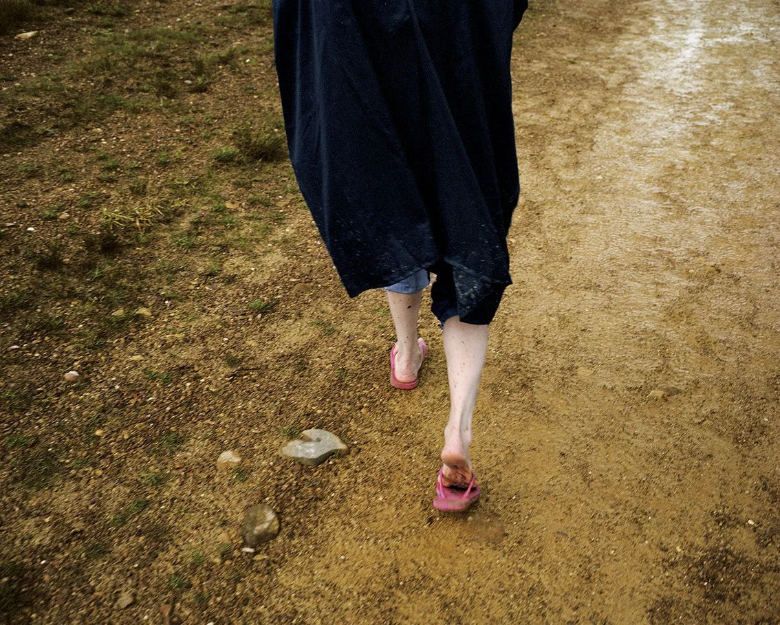 A person walking barefoot on a dirt path, wearing pants and a long coat, with pink sandals instead of shoes.